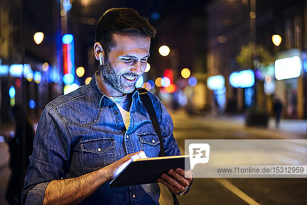 Lächelnder Mann mit drahtlosen Kopfhörern mit digitalem Tablet in der Stadt bei Nacht