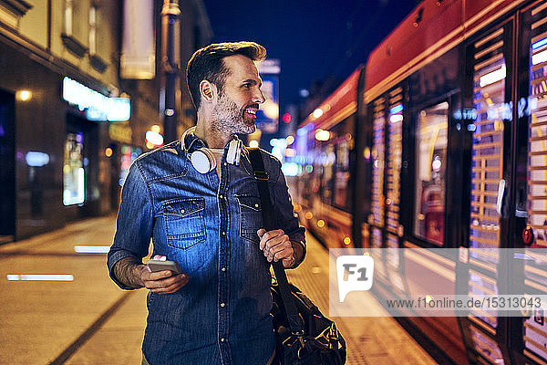 Smiling man using his smartphone in the city at night while waiting for the tram