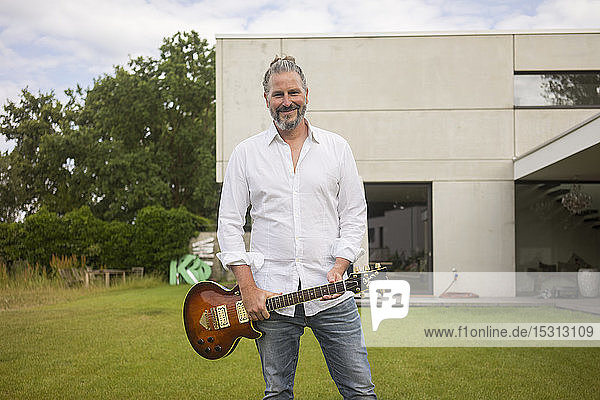 Portrait of mature man standing on lawn in front of his house holding guitar