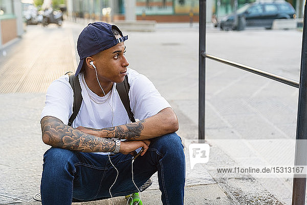 Tätowierter junger Mann sitzt auf seinem Skateboard und hört mit Smartphone und Kopfhörern Musik
