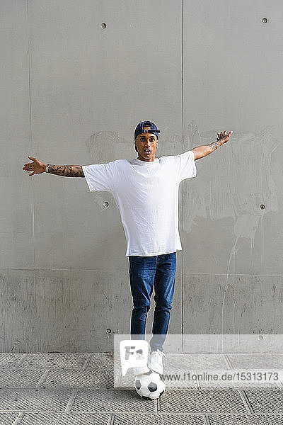 Portrait of tattooed young man balancing on football in front of concrete wall