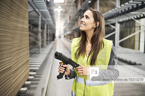 Lächelnde Frau in Fabrikhalle mit Sicherheitsweste und Barcode-Scanner