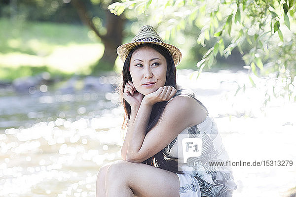 Junge Frau  die einen Strohhut trägt und am Flussufer sitzt