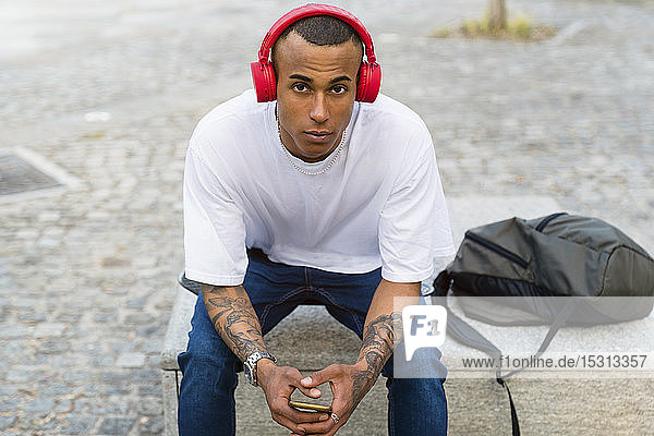 Porträt eines tätowierten jungen Mannes  der auf einer Bank sitzt und mit Smartphone und roten Kopfhörern Musik hört