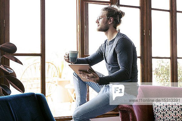 Nachdenklicher junger Mann zu Hause mit einem digitalen Tablet  der aus dem Fenster schaut