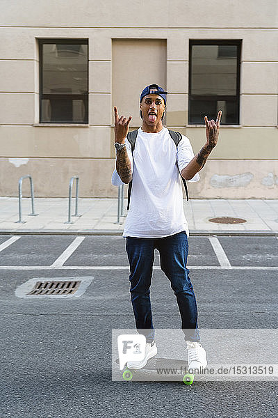 Tätowierter junger Mann steht auf Skateboard  streckt die Zunge heraus und zeigt Rock-and-Roll-Zeichen