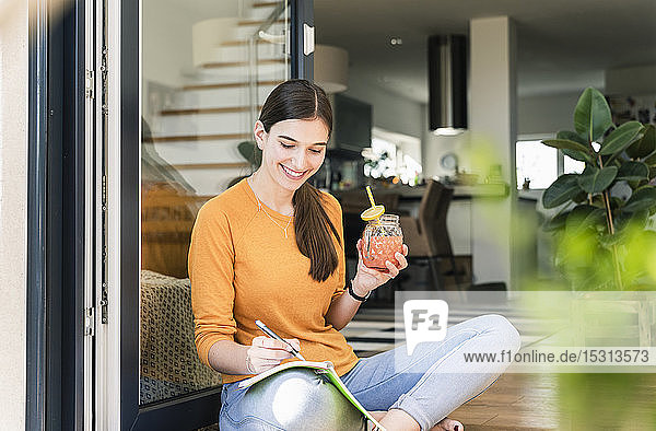 Junge Frau sitzt mit gesundem Getränk und Notizbuch an der Terrassentür