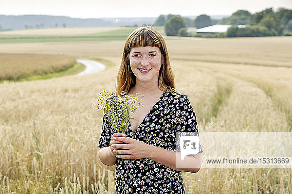 Porträt einer lächelnden jungen Frau mit Kamillenblütenstrauss vor einem Getreidefeld stehend