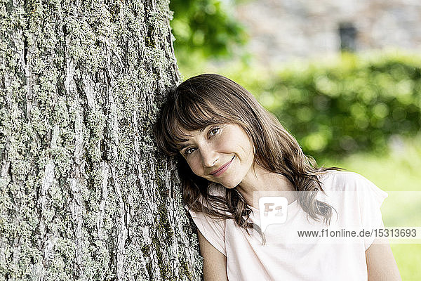 Bildnis einer lächelnden brünetten Frau  die an einen Baumstamm gelehnt ist