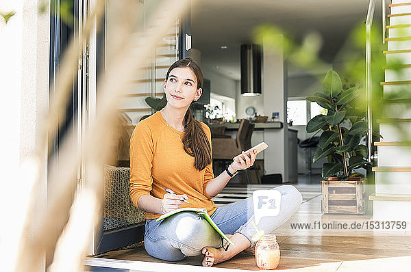 Junge Frau sitzt mit Handy und Notebook an der Terrassentür