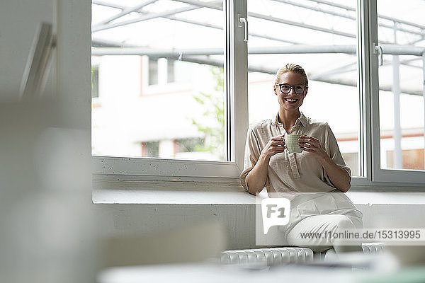 Porträt einer lächelnden jungen Geschäftsfrau mit Tasse Kaffee am Fenster sitzend