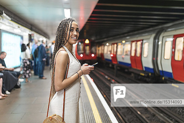 Porträt einer lächelnden jungen Frau mit Smartphone  die am Bahnsteig einer U-Bahn-Station wartet  London  Großbritannien