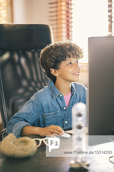 Lächelnder Junge sitzt zu Hause am Schreibtisch und benutzt einen PC