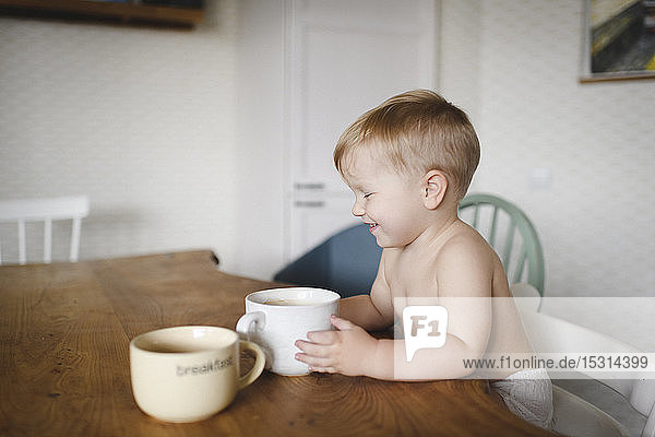 Laughing little boy sitting at kitchen table holding cup