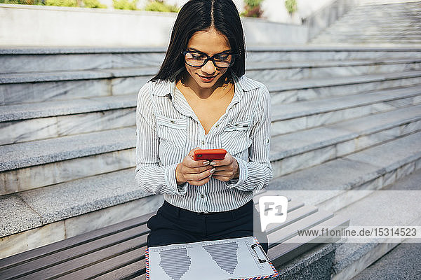 Junge Geschäftsfrau  die auf einer Bank sitzt und ihr Handy überprüft