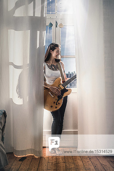 Glückliche junge Frau  die zu Hause mit einer Gitarre am Fenster steht