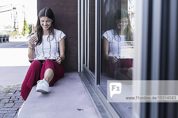 Junge Frau sitzt auf der Fensterbank eines Gebäudes und benutzt ein Tablett