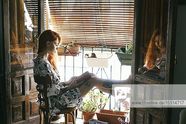 Rothaarige tätowierte Frau zu Hause beim Lesen eines Buches am Fenster