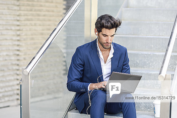 Businessman wearing earphones using laptop outdoors in the city
