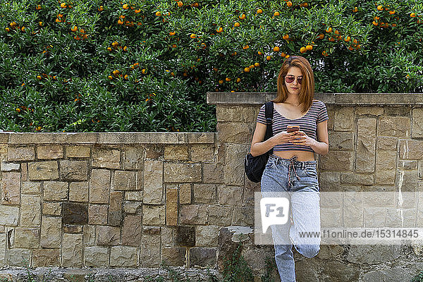 Woman with backpack leaning against stone wall using cell phone