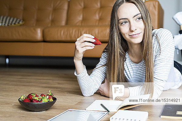 Entspannte junge Frau liegt zu Hause auf dem Boden und isst Erdbeeren