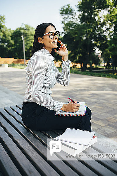 Junge Geschäftsfrau sitzt auf einer Bank  telefoniert mit dem Handy und macht sich Notizen