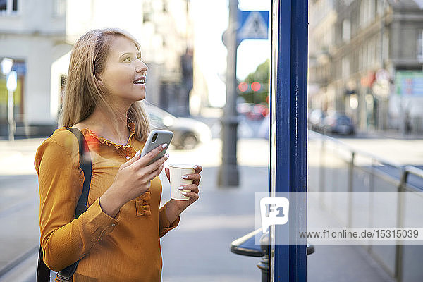Smiling young woman with smartphone and takeaway coffee checking the schedule at bus stop