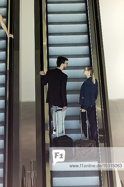 Zwei junge Geschäftspartner mit Gepäck unterhalten sich auf einer Rolltreppe