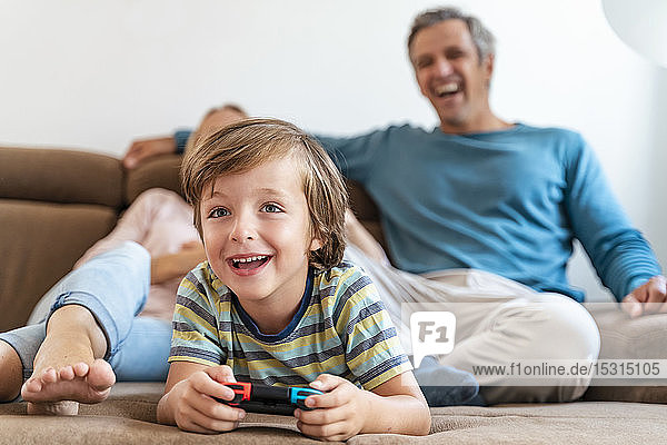 Boy lying on couch at home playing video game with parents watching