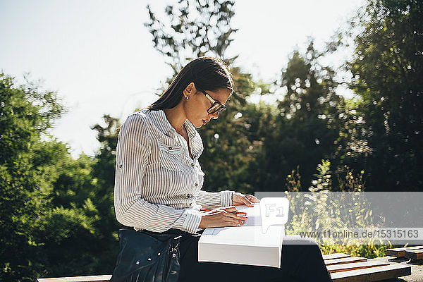 Junge Frau sitzt auf einer Parkbank und betrachtet ein geöffnetes Buch