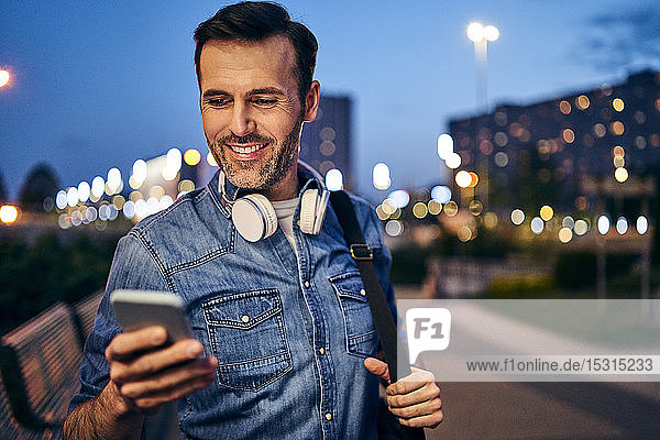 Lächelnder Mann benutzt abends in der Stadt sein Smartphone