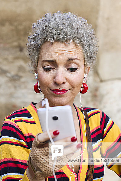 Porträt einer gepiercten reifen Frau mit grauem lockigem Haar beim Blick auf ein Mobiltelefon