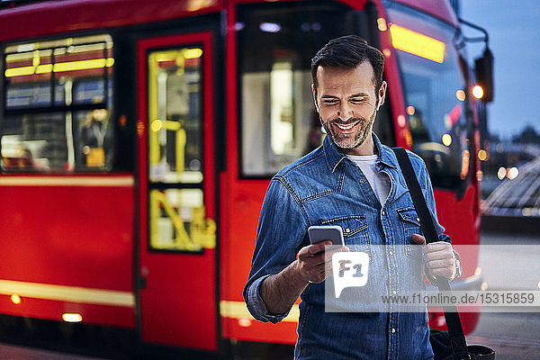Lächelnder Mann mit Smartphone in der Stadt mit Straßenbahnfahrten im Hintergrund