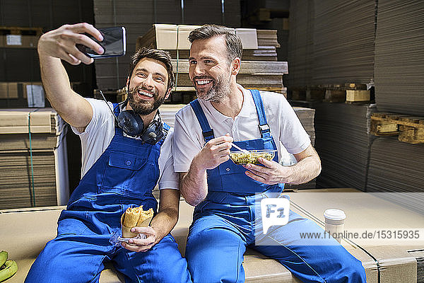 Two happy workers having lunch break and taking a selfie in factory