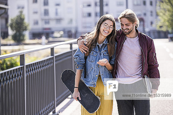Glückliches junges Paar mit Skateboard und Smartphone auf einer Brücke
