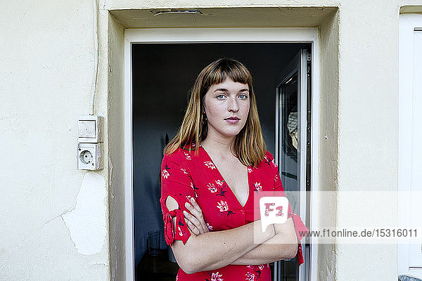 Porträt einer ernsten jungen Frau mit verschränkten Armen vor geöffneter Balkontür stehend
