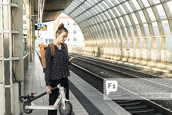 Junge Frau mit Elektroroller und Handy am Bahnhof