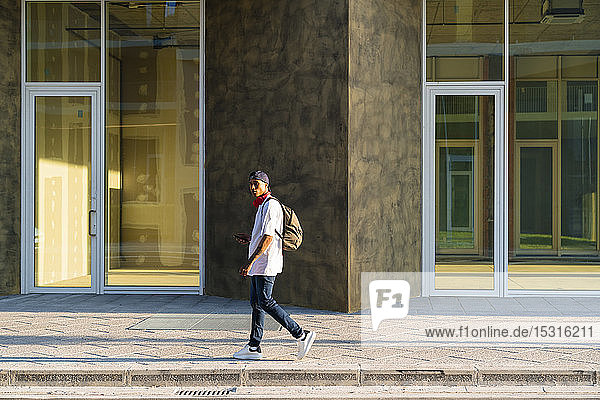 Junger Mann mit Rucksack geht bei Sonnenlicht auf dem Bürgersteig
