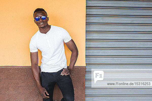 Porträt eines jungen Mannes mit weißem T-Shirt und Sonnenbrille  der an einer Wand lehnt