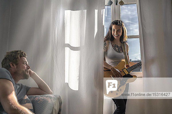 Mann schaut junge Frau zu Hause am Fenster mit einer Gitarre an