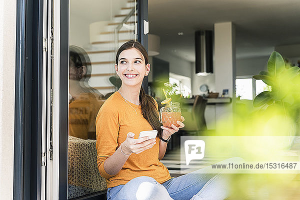 Lächelnde junge Frau sitzt mit gesundem Getränk und Handy an der Terrassentür