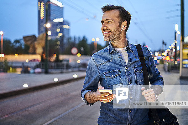 Smiling man using smartphone and listening to music through wireless headphones while waiting at tram stop during evening commute after work