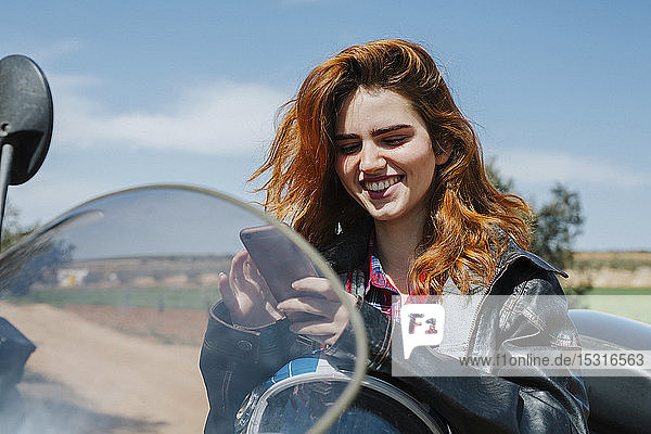 Porträt einer glücklichen rothaarigen Frau auf Motorrad mit Blick auf Handy