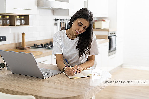 Young woman with laptop taking notes at home