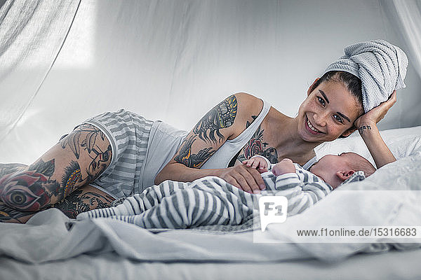 Porträt einer lächelnden tätowierten jungen Frau mit ihrem Baby im Himmelbett