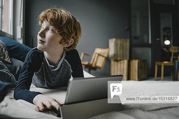 Rothaariger Junge auf Couch liegend mit digitalem Tablett aus dem Fenster schauend
