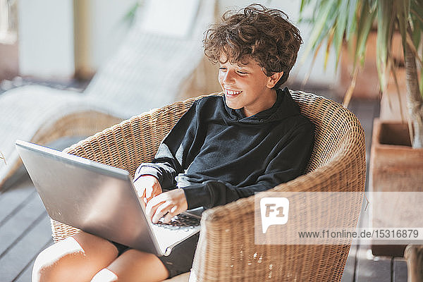 Lächelnder Junge sitzt mit Laptop auf einem Korbstuhl