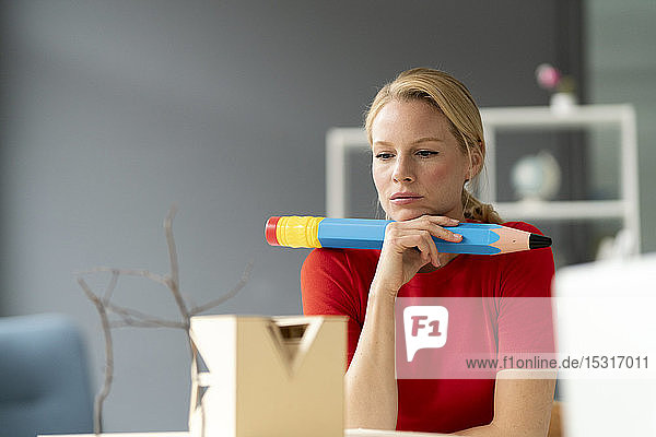 Junge Frau im Büro mit übergroßem Stift und Architekturmodell auf dem Schreibtisch