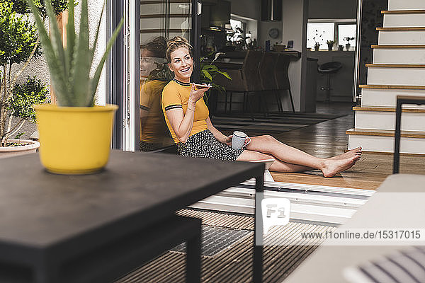 Entspannte Frau sitzt zu Hause an der Terrassentür und benutzt ein Smartphone