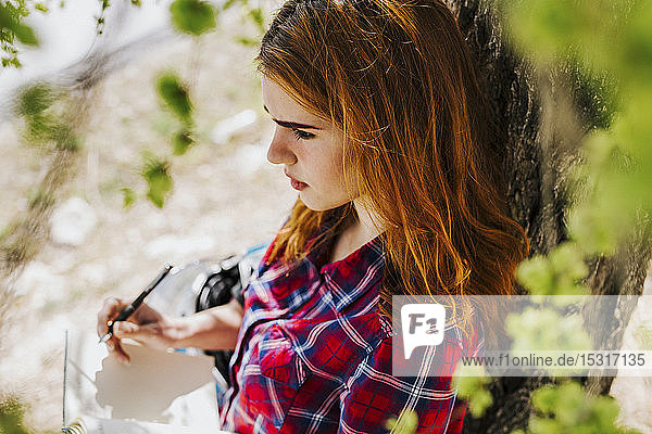 Pensive redheaded woman leaning against tree trunk taking notes